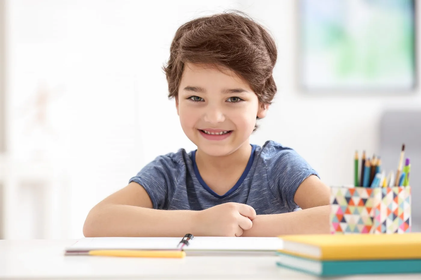 A smiling young boy sitting at a desk with an open notebook, colored pencils, and books.